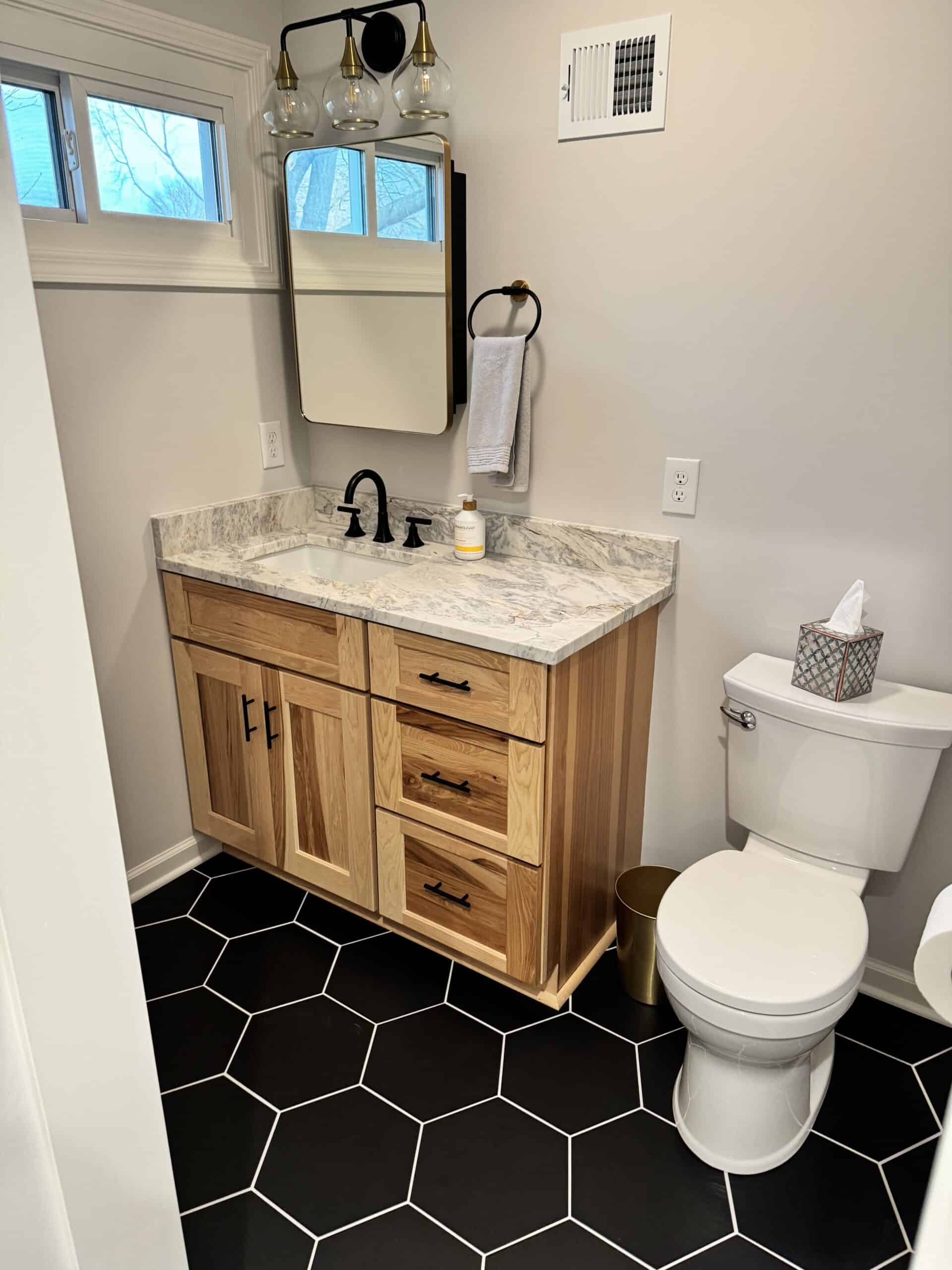 Single-sink vanity with light wood cabine Single-sink vanity with light wood cabinet and marble countertop beside a toilet, on black hexagonal tile flooring